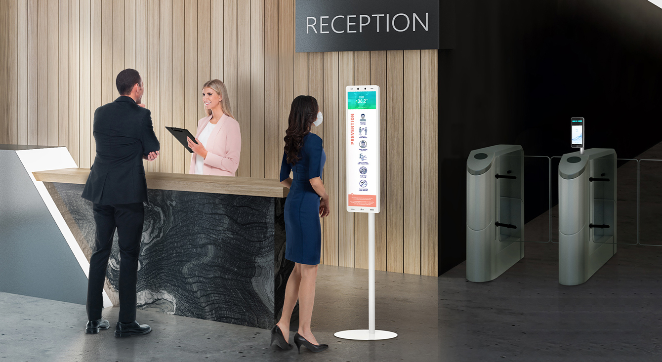 The 29" Thermal sensing terminal is placed near the reception desk. And The employee with a mask stands in front of the terminal to check her fever .