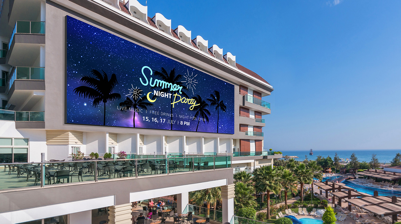 A large LED is installed on the building’s outer wall next to a resort’s outdoor swimming pool by the beach, and it clearly shows the resort’s event advertisement.
