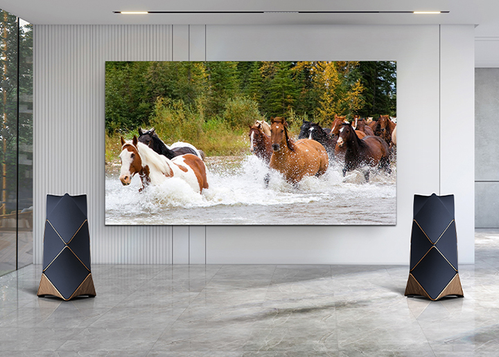 An LG MAGNIT installed on a living room wall, is vividly displays a herd of horses galloping through the water and dynamic sound coming from the Beolab 90 speakers located on both sides of the LG MAGNIT.